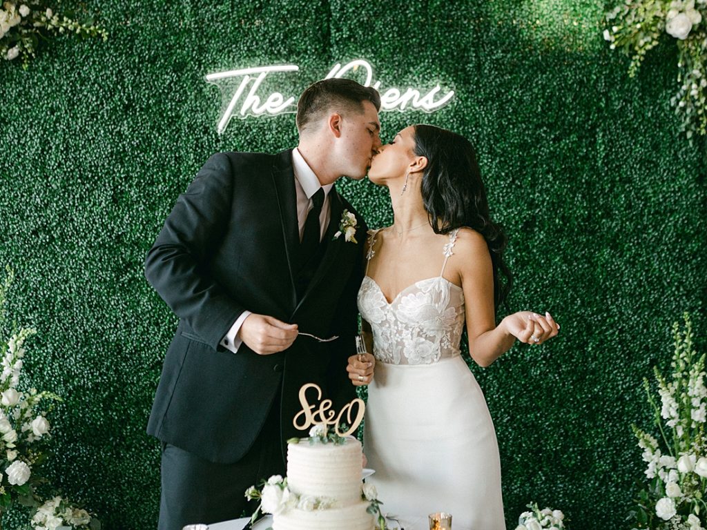 Bride and groom kiss after cutting the cake with a hedge background and neon sign