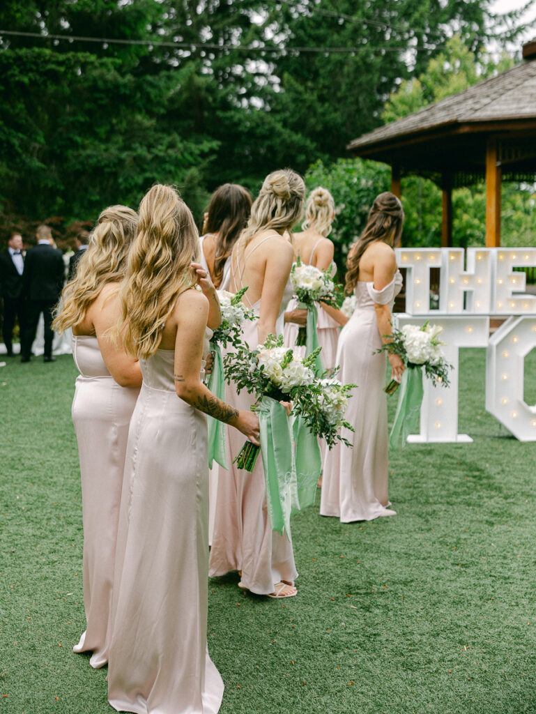 Emily and her bridesmaids laugh together holding white spring bouquets with sage green ribbons.