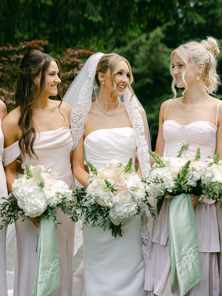Emily and her bridesmaids laugh together holding white spring bouquets with sage green ribbons.