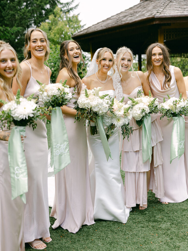Emily and her bridesmaids laugh together holding white spring bouquets with sage green ribbons.