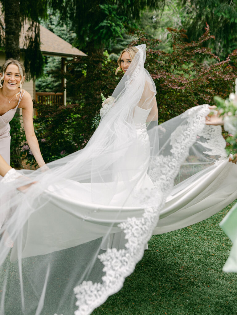 Bride Emily smiles as her bridesmaids spread her long lace-trimmed veil during outdoor wedding portraits.
