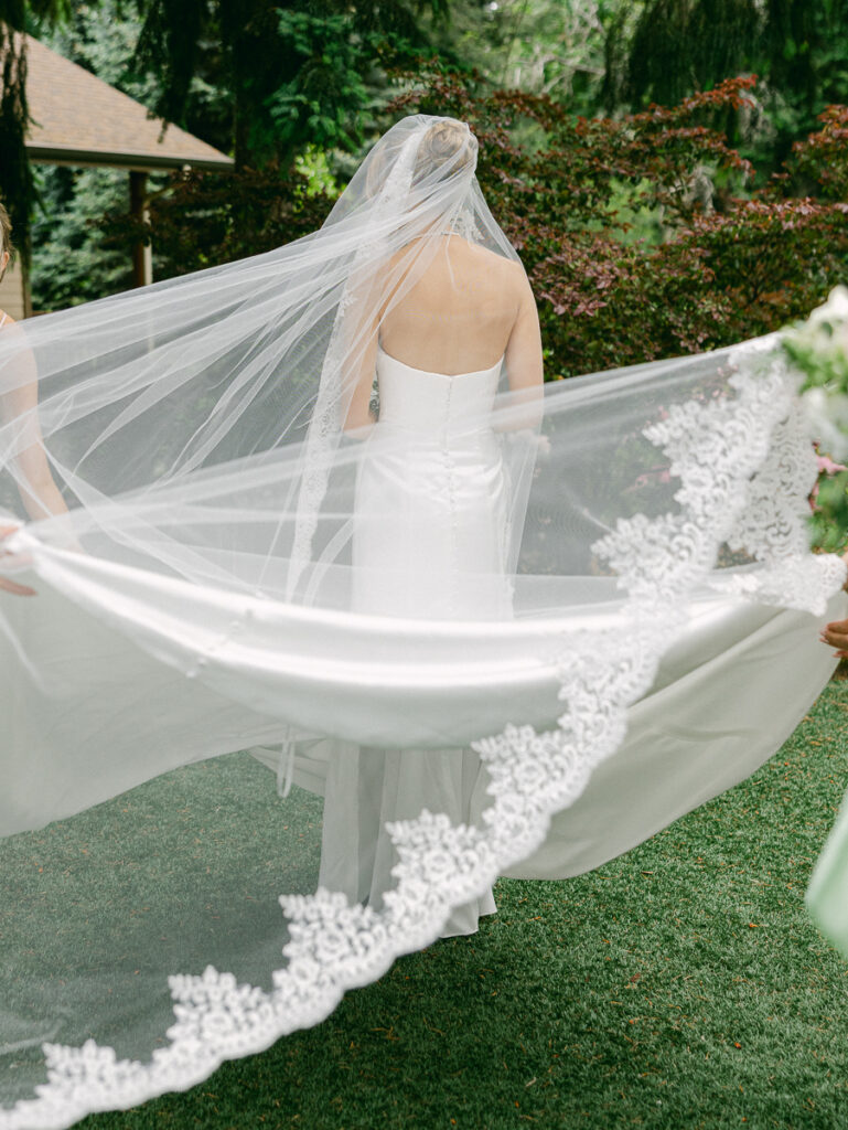 Bride Emily smiles as her bridesmaids spread her long lace-trimmed veil during outdoor wedding portraits.