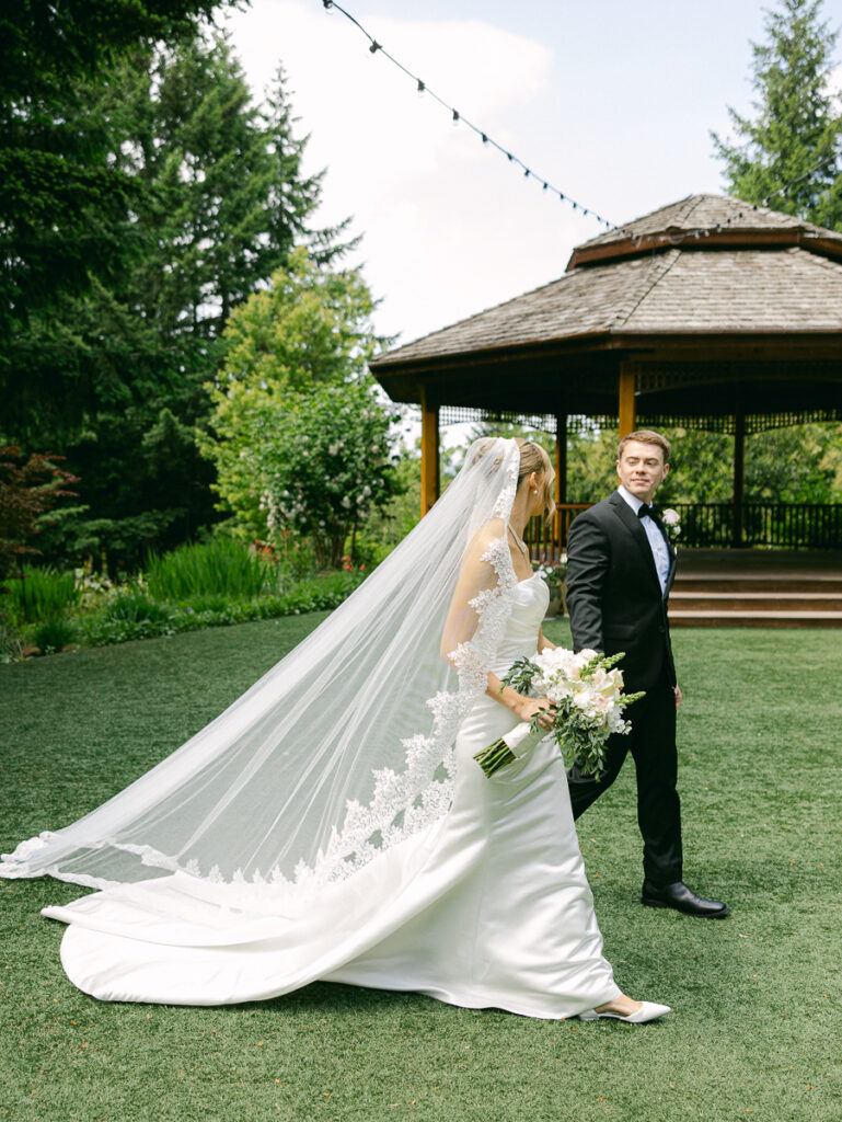 Owen and Emily walk together outdoors with her long lace wedding veil flowing behind them.