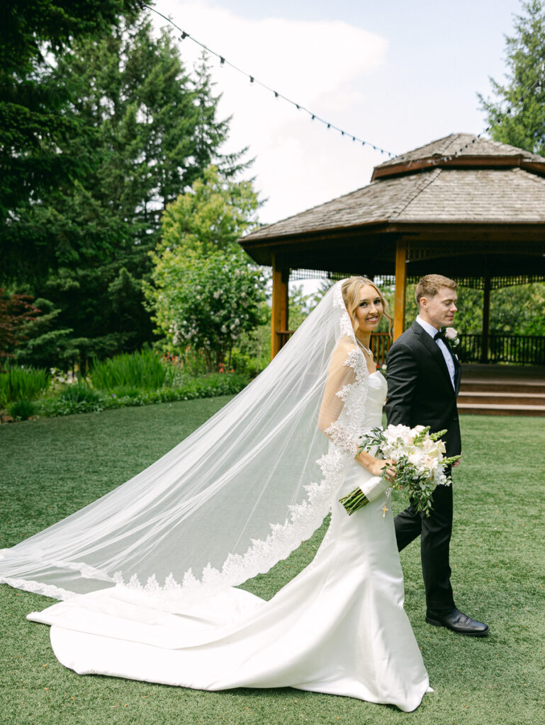 Owen and Emily walk together outdoors with her long lace wedding veil flowing behind them.
