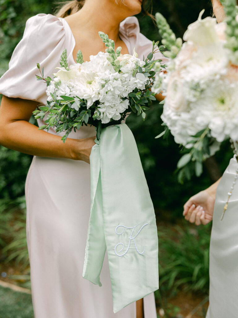 Emily and her bridesmaids laugh together holding white spring bouquets with sage green ribbons.